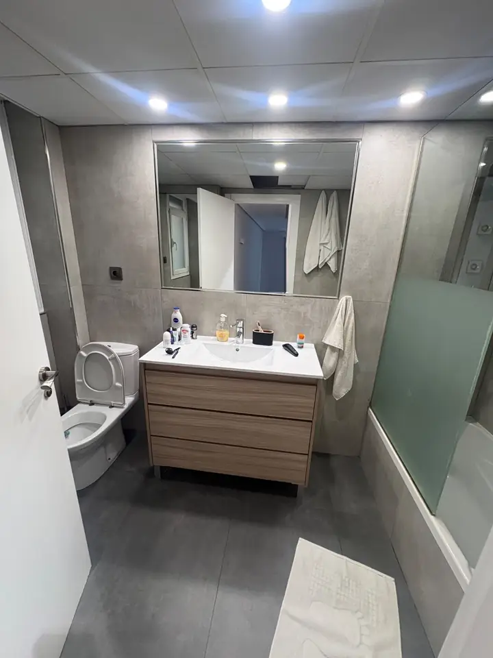 Foto real de un baño antes de la remodelación: lavamanos con mueble de madera, WC, tina con mampara y cerámica gris
